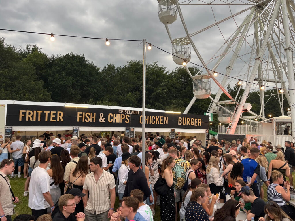 Stor menneskemængde foran madbod med skilte for ‘Fritter’, ‘Fish & Chips’, ‘Chicken’ og ‘Burger’ på en udendørs festival med pariserhjul i baggrunden.