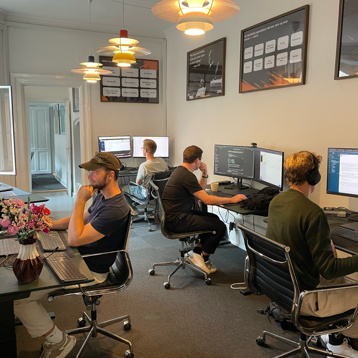 Fire medarbejdere arbejder ved hver deres computer i et lyst kontor med plakater på væggen